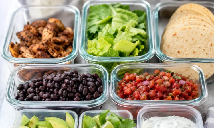 Healthy taco meal prep containers with chicken, tortillas, lettuce, beans, avocado, salsa, cilantro, lime, and yogurt sauce