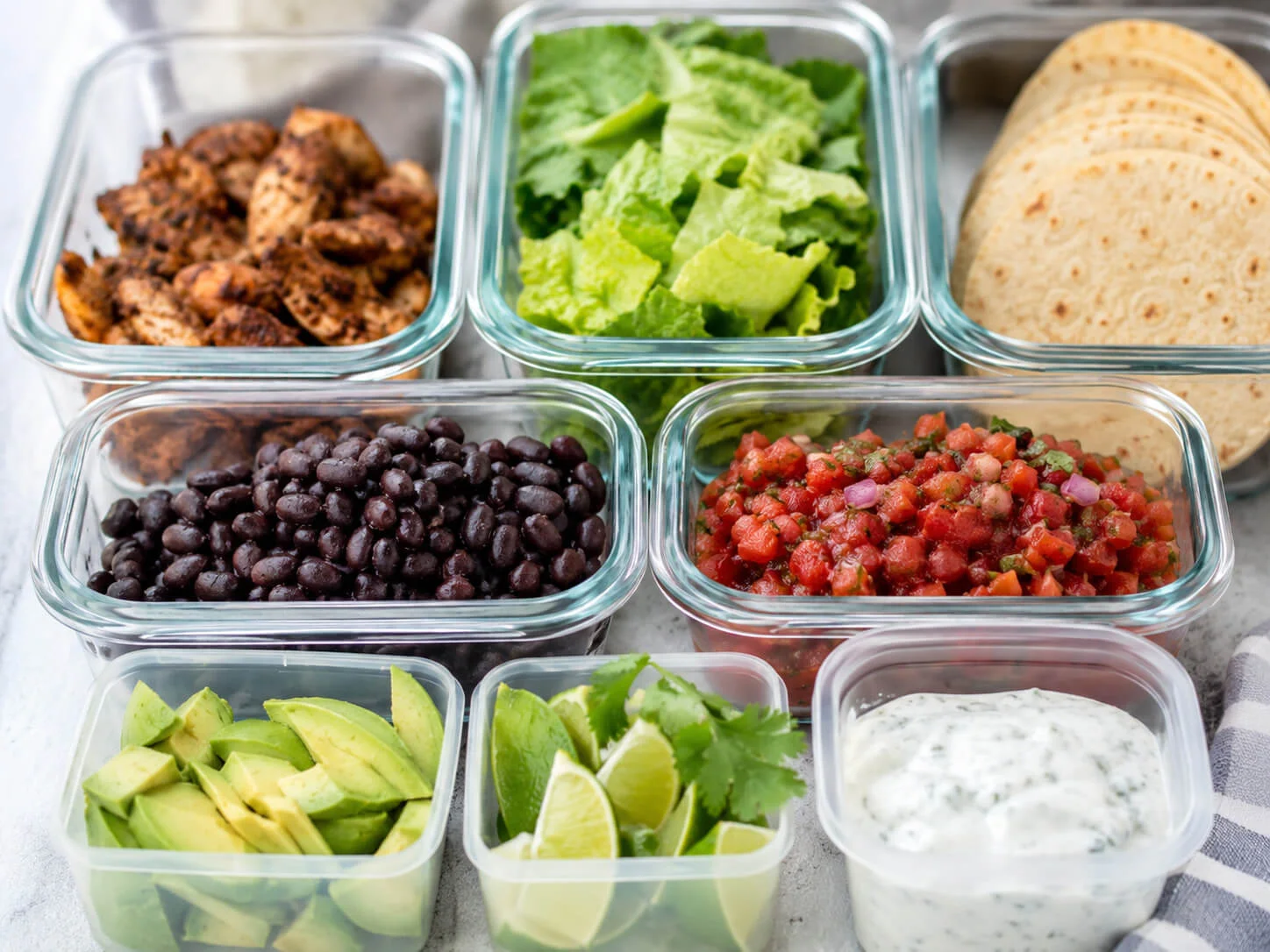 Healthy taco meal prep containers with chicken, tortillas, lettuce, beans, avocado, salsa, cilantro, lime, and yogurt sauce