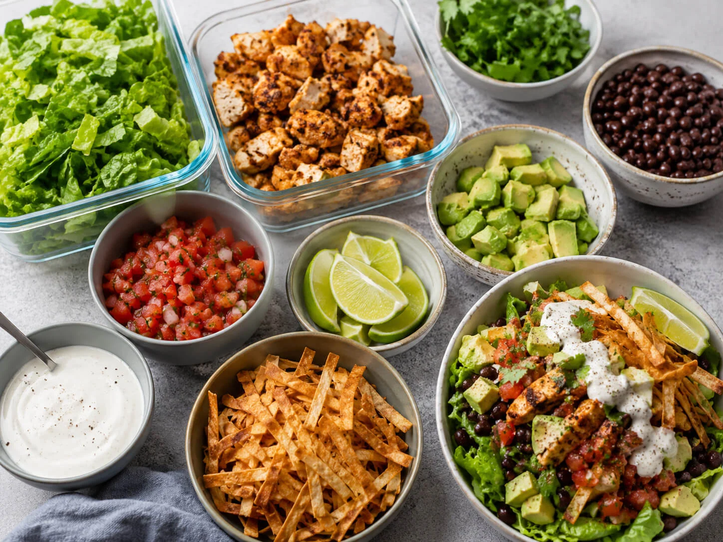 Healthy taco salad meal prep spread with chicken, lettuce, black beans, avocado, salsa, cilantro, lime, tortilla strips, and yogurt dressing